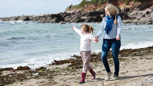 A woman and young girl walk along the shoreline in warm clothes, rocks in the background